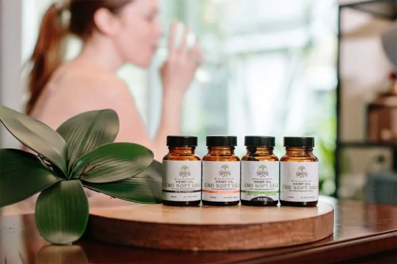 Four bottles of CBD soft gels are placed on a wooden surface beside a green plant. In the blurred background, reminiscent of a guest relaxing in one of the finest hotels in London, a person is drinking a glass of water. The image suggests a natural and wellness-focused setting.