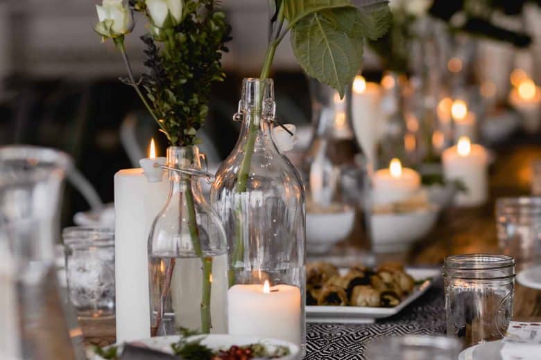 A dining table is elegantly set with decorative clear glass bottles holding white flowers, white candles, and flickering tea lights. Plates with food are arranged on the table, creating a warm and inviting ambiance reminiscent of the finest hotels in London. The soft lighting enhances the intimate setting.