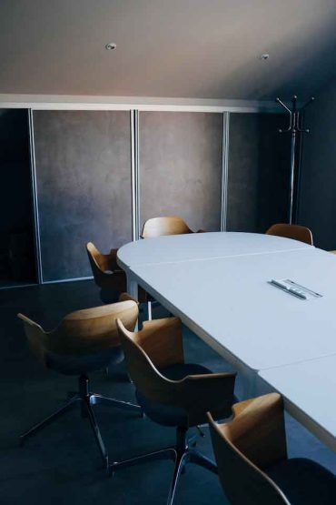 A modern conference room with a large white oval table surrounded by several wooden swivel chairs. The room has a minimalistic design with a grey wall in the background and a coat rack in the corner. A set of documents lies on the table, reminiscent of meeting spaces offered by top hotels in London.