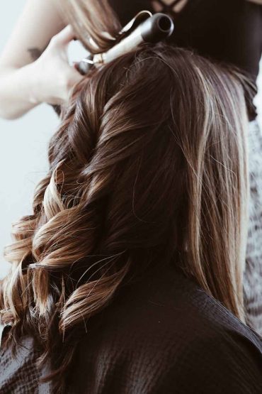 A stylist, perhaps at one of the luxurious hotels in London, uses a curling iron to create loose curls in a woman's long brown hair. The scene focuses on the back of the woman's head as the stylist works diligently.