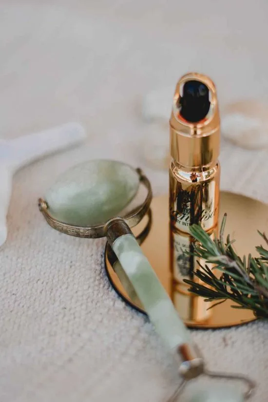 A jade-handled facial roller and a golden skincare bottle with a pump dispenser rest elegantly on a round mirror. A sprig of rosemary adds a touch of nature, all set against a soft, beige fabric background that evokes the luxury of hotels in London.