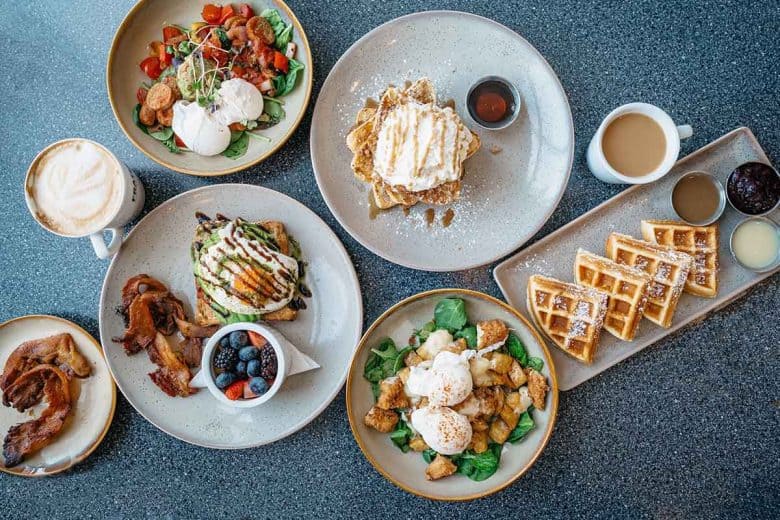 A top view of a breakfast spread at one of the finest hotels in London on a gray table, including various dishes: waffles with syrup, eggs benedict on greens and potatoes, fresh berries, a plate with bacon, toast with toppings, a latte, and a cup of coffee. Each dish is served on white plates.