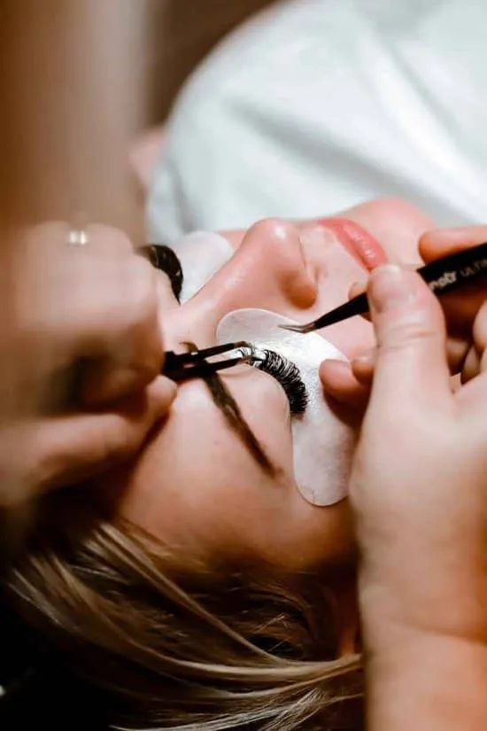 A person is lying down with their eyes closed, receiving eyelash extensions. In a serene room that could rival the finest hotels in London, a technician uses tweezers to apply the extensions while under-eye pads rest beneath the person's eyes, highlighting the meticulous process and focus on detail.