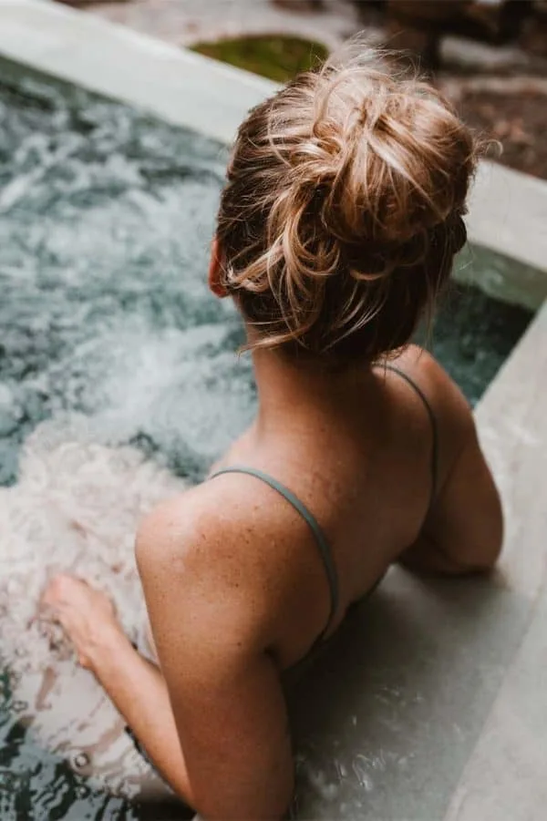 A woman with blonde hair in a messy bun is relaxing in a hot tub at one of the luxurious hotels in London. She is seen from behind, wearing a grey strappy top. The water around her is bubbly, and the edge of the hot tub is visible. The setting appears calm and serene.