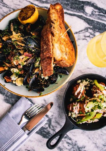 A top-down view of a meal with a bowl of mussels and lemon, garnished with herbs and served with toasted bread. Next to it, a small skillet contains a dish topped with greens. A knife and fork rest on a napkin, and a yellow drink is nearby, reminiscent of fine dining experiences often found in hotels in London.