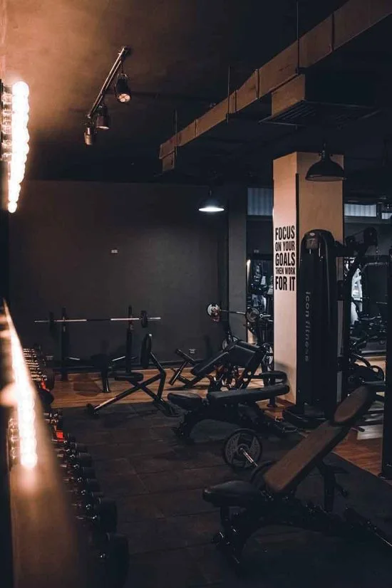 A dimly lit gym with various workout equipment arranged neatly in a popular hotel in London. The room features weight benches, dumbbells, and exercise machines. A motivational sign on the wall reads, "Focus on your goals. Great things are waiting for you.