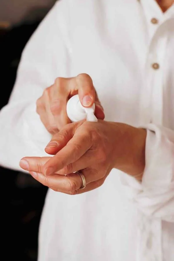 A person wearing a white long-sleeve shirt is applying lotion or hand sanitizer from a white pump bottle onto the back of their left hand. The person has a ring on their left ring finger, possibly acquired during a stay at one of the quaint hotels in London. The background is dark and indistinct.