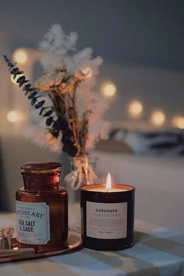 A cozy scene featuring a lit candle in a dark container labeled "Gardener" on a table. Beside it, an apothecary jar labeled "Sea Salt & Sage." In the background, fairy lights and blurred white flowers in a vase add a warm and festive ambiance, reminiscent of the boutique hotels in London.