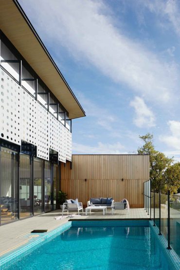 A modern home with large windows and perforated white panels on the upper level is adjacent to a rectangular swimming pool, reminiscent of luxury hotels in London. The outdoor space features a wooden deck with white lounge furniture. The sky is partly cloudy, and greenery is visible in the background.