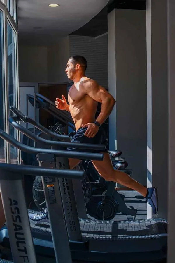 A muscular man is running on a treadmill in a gym with large windows, allowing natural light to pour in. He is shirtless and wears black shorts and sneakers, focusing intently on his workout. The background includes other exercise equipment, reminiscent of the sleek fitness centers found in top hotels in London.