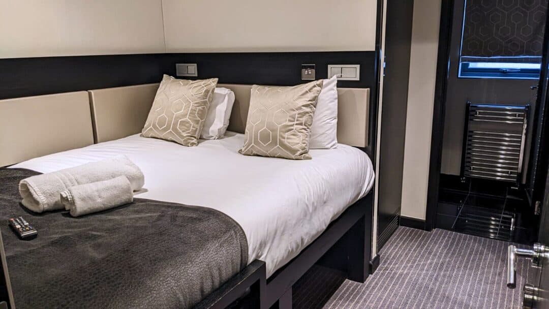 A neatly made bed with two patterned pillows and folded white towels in a modern hotel room. A TV remote rests on the bed, and the room features neutral colors, a window, and a small radiator in the background.