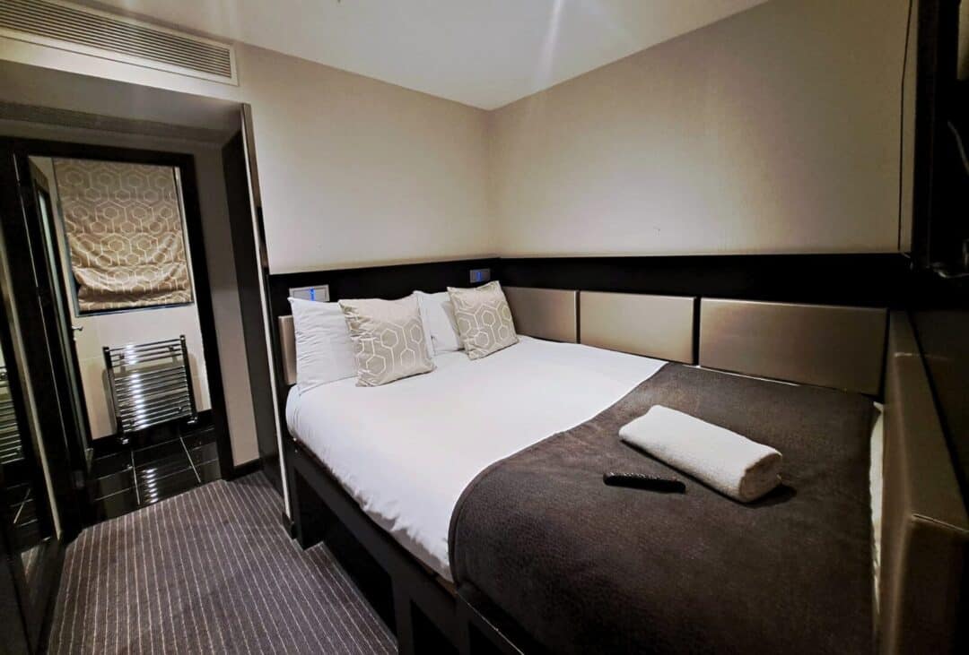 A modern hotel room with a neatly made double bed, three decorative pillows, a folded towel, and a TV remote on a dark blanket. There is a doorway leading to a bathroom with a towel radiator and patterned window shade.