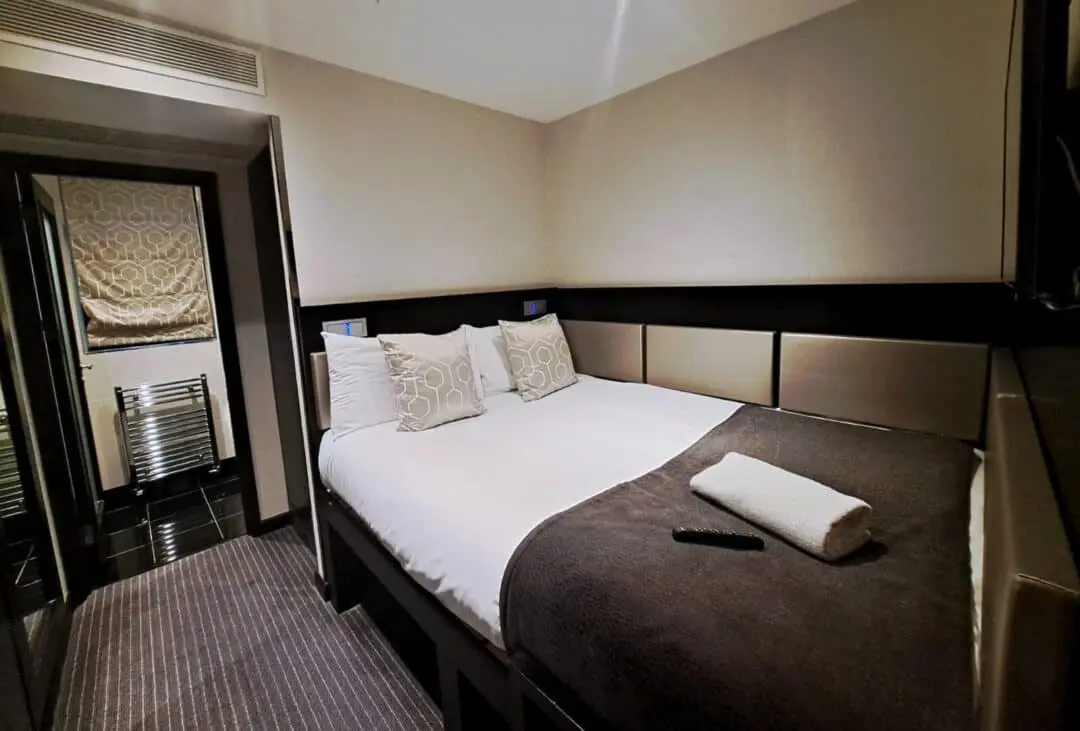 A modern hotel room with a neatly made double bed, three decorative pillows, a folded towel, and a TV remote on a dark blanket. There is a doorway leading to a bathroom with a towel radiator and patterned window shade.
