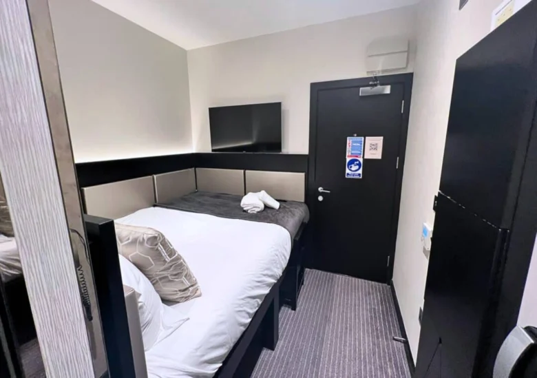 A small, modern hotel room with a neatly made bed, white and grey bedding, a wall-mounted TV, and a black door. The room has minimal decor and compact furniture, maximizing the limited space.