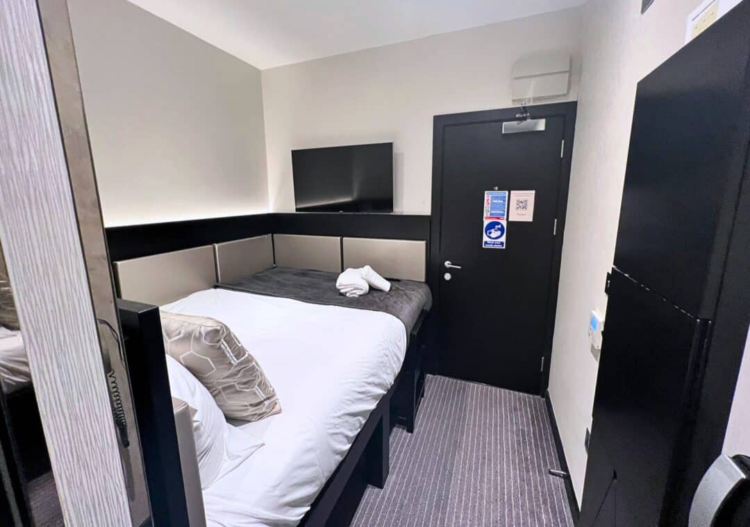 A small, modern hotel room with a neatly made bed, white and grey bedding, a wall-mounted TV, and a black door. The room has minimal decor and compact furniture, maximizing the limited space.
