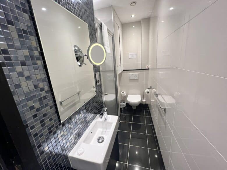 A modern, narrow bathroom with glossy black floor tiles, white wall tiles, a small sink with a mirror, a wall-mounted toilet, and a walk-in shower with a glass door. The decor includes mosaic tiles and bright lighting.