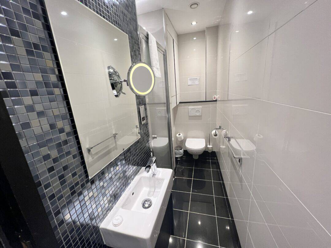 A modern, narrow bathroom with glossy black floor tiles, white wall tiles, a small sink with a mirror, a wall-mounted toilet, and a walk-in shower with a glass door. The decor includes mosaic tiles and bright lighting.