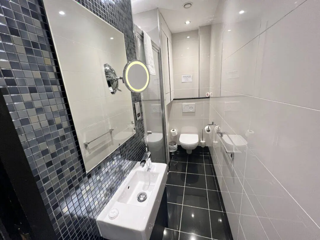 A modern, narrow bathroom with glossy black floor tiles, white wall tiles, a small sink with a mirror, a wall-mounted toilet, and a walk-in shower with a glass door. The decor includes mosaic tiles and bright lighting.