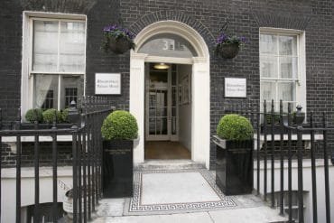 Entrance of Bloomsbury Palace Hotel, a standout among hotels in London, featuring a black brick façade, white arched doorway with the number 31 above, and two well-maintained green shrubs in black planters flanking the entry. Iron railings, hanging flower baskets, and two windows are visible.