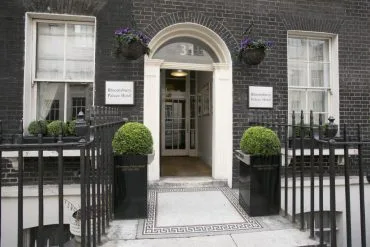 Entrance of Bloomsbury Palace Hotel, a standout among hotels in London, featuring a black brick façade, white arched doorway with the number 31 above, and two well-maintained green shrubs in black planters flanking the entry. Iron railings, hanging flower baskets, and two windows are visible.