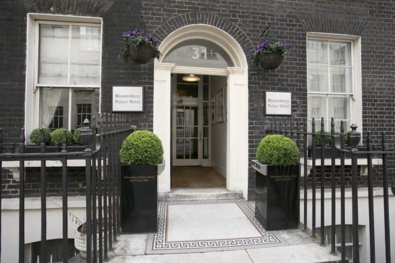 Entrance of Bloomsbury Palace Hotel, a standout among hotels in London, featuring a black brick façade, white arched doorway with the number 31 above, and two well-maintained green shrubs in black planters flanking the entry. Iron railings, hanging flower baskets, and two windows are visible.