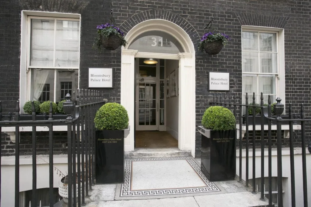 Entrance of Bloomsbury Palace Hotel, a standout among hotels in London, featuring a black brick façade, white arched doorway with the number 31 above, and two well-maintained green shrubs in black planters flanking the entry. Iron railings, hanging flower baskets, and two windows are visible.