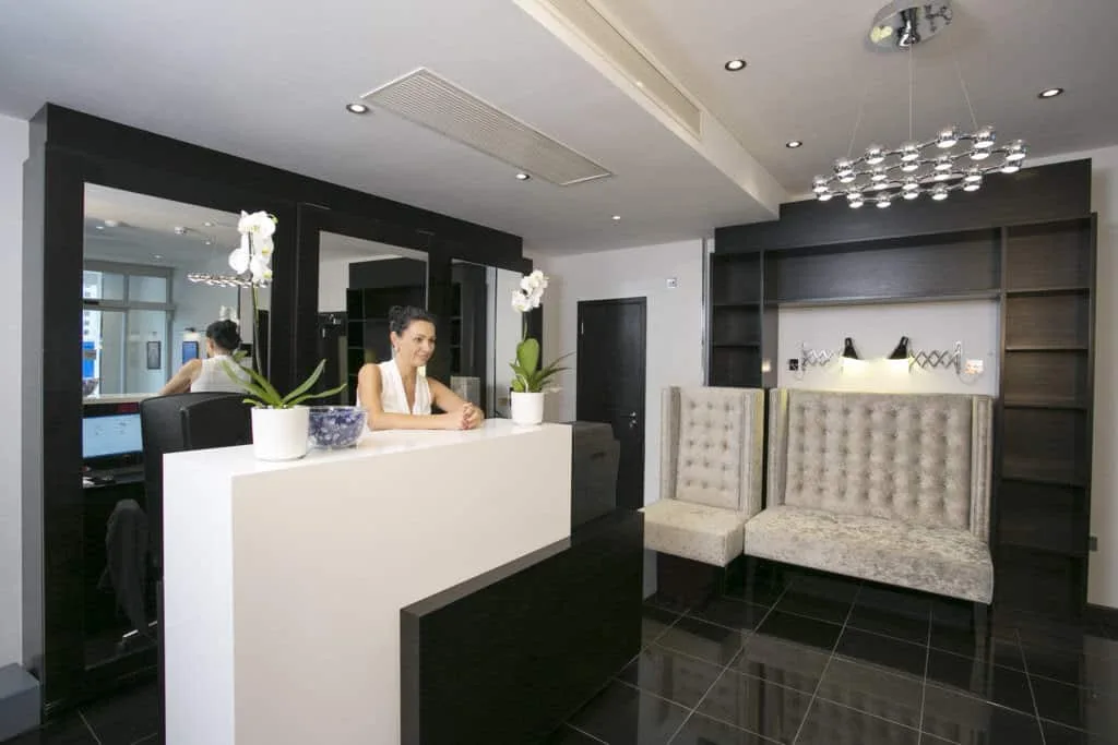 A modern reception area with a woman standing behind a white desk. The room features dark wood accents, tufted beige seating, potted plants, and contemporary lighting. Shelving units and mirrors are in the background, and the floor is tiled in black—reminiscent of the best hotels in London.