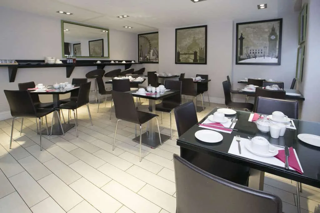 A modern dining area in one of the finest hotels in London features black chairs and tables set with white dishes, cups, and red napkins. The room showcases large framed pictures on the walls and a shelf holding condiments. The floor is tiled in a light color, and the space is well-lit.