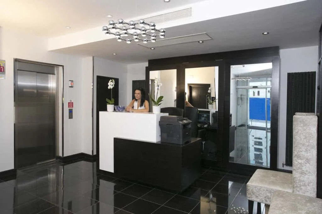 A modern reception area with a shiny black tiled floor greets guests at one of the finest hotels in London. A woman is seated behind a sleek black and white reception desk. To the left is an elevator with silver doors, and a stylish, contemporary chandelier hangs from the ceiling, casting reflections on both floor and glass.