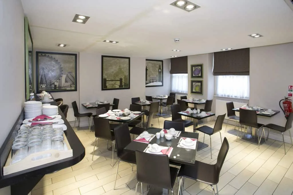 A modern dining area with several neatly arranged tables, each set with plates, cups, and red napkins. The walls are adorned with framed black and white cityscape artwork reminiscent of hotels in London. A table on the left holds additional dishes and glasses. Windows have brown Roman shades.