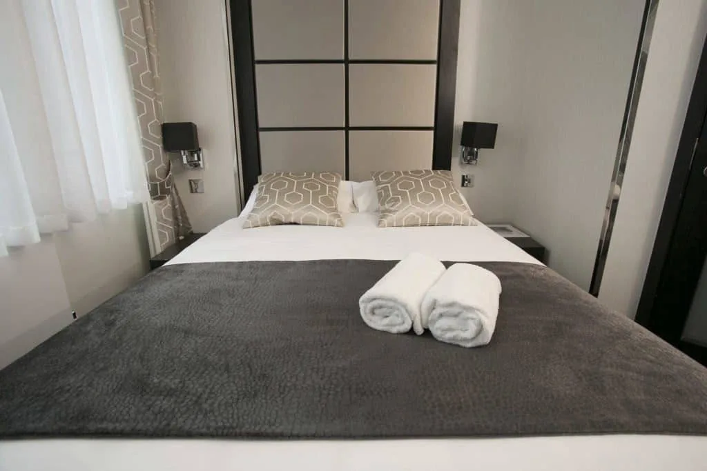 A neatly made bed with a dark grey blanket and two patterned pillows is adorned with two rolled white towels placed in the center. Evoking the elegance of hotels in London, the room is decorated in a modern style with light grey walls, white curtains, and black wall-mounted lamps.