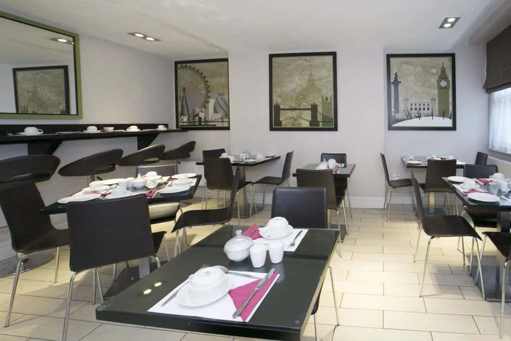 A modern, clean café with black tables and chairs, reminiscent of stylish hotels in London. The tables are set with white dishes, cups, and folded pink napkins. The walls are adorned with framed cityscape artwork, and mirrors provide a spacious feel. The space is well-lit with natural and artificial light.
