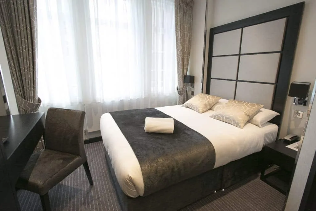 A neatly arranged hotel room in one of the charming hotels in London, featuring a double bed with white and gray linens, two patterned pillows, and a neatly folded towel on top. The room has large windows with curtains, a dark wooden chair, and a small desk beside the bed.