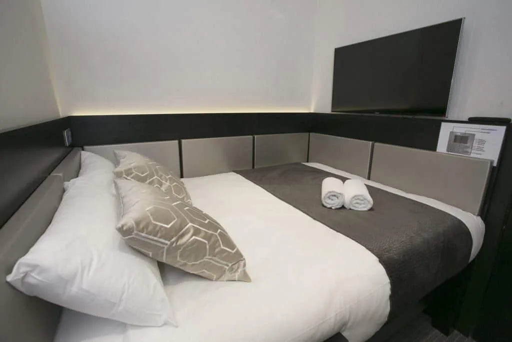 A sleek, modern bedroom in one of the finest hotels in London features a neatly made bed with white and grey bedding, two patterned pillows, and two rolled towels placed on the bed. A wall-mounted flat-screen TV is visible at the foot of the bed, and the room has minimalist decor.