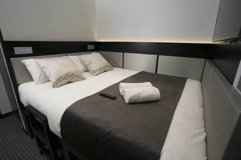 A modern, compact hotel room in one of the finest hotels in London features a neatly made double bed with white sheets, a dark gray blanket, and two beige patterned pillows. Two folded white towels and a TV remote control lie on the bed. The room has minimalistic decor with clean lines and soft lighting.