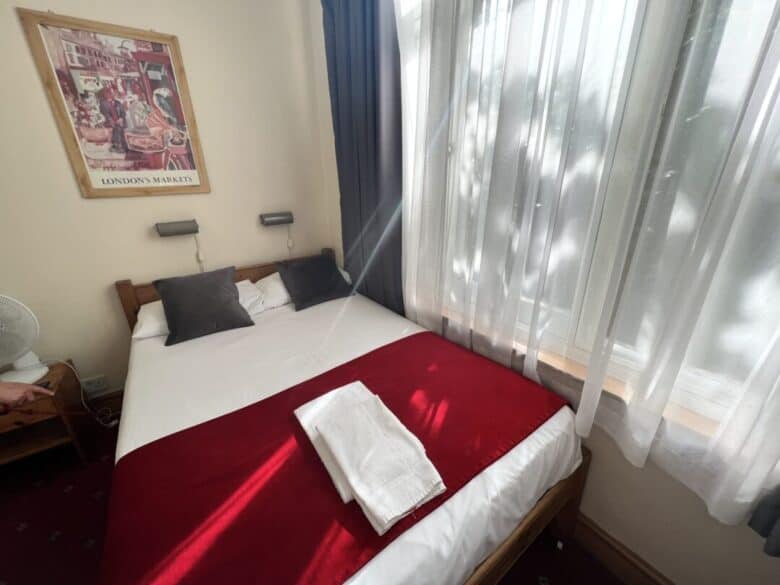 A cozy bedroom with a double bed, red blanket, and two white towels. Sunlight streams through sheer white curtains covering a large window. A London-themed poster and two pillows are on the wall above the bed.