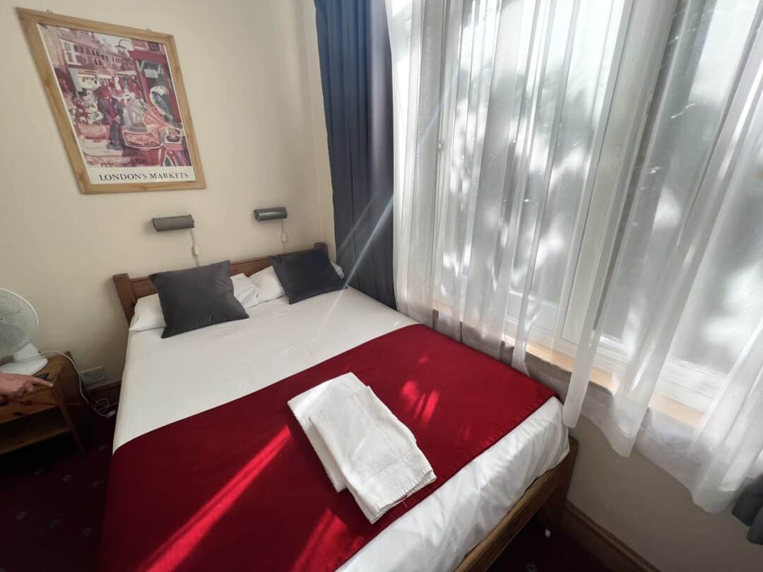A cozy bedroom with a double bed, red blanket, and two white towels. Sunlight streams through sheer white curtains covering a large window. A London-themed poster and two pillows are on the wall above the bed.