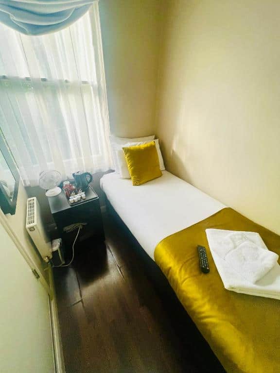 A small, cozy bedroom with a single bed, gold and white bedding, and a remote and folded towels on top. A bedside table holds a fan, kettle, cups, and snacks, next to a window with sheer curtains.