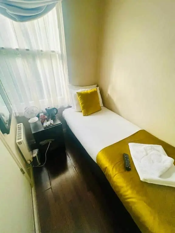 A small, cozy bedroom with a single bed, gold and white bedding, and a remote and folded towels on top. A bedside table holds a fan, kettle, cups, and snacks, next to a window with sheer curtains.