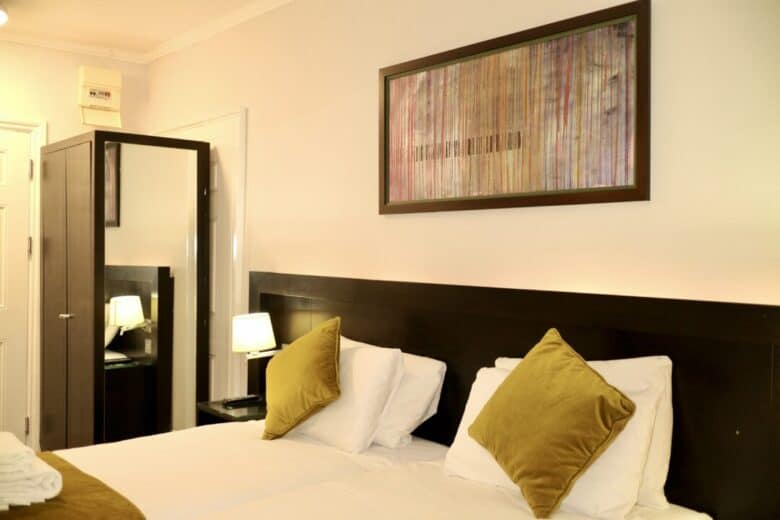 A neatly made bed with two green pillows, a dark wooden headboard, and a matching wardrobe with a mirror. A lit bedside lamp and framed abstract art hang on the wall above the bed.
