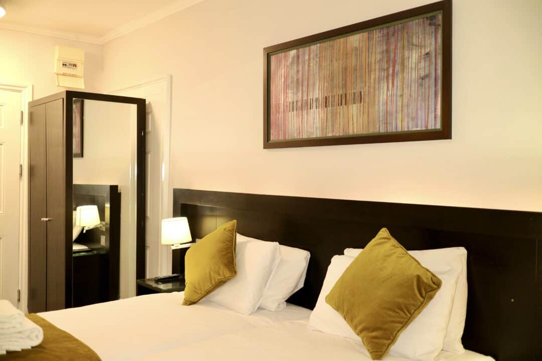A neatly made bed with two green pillows, a dark wooden headboard, and a matching wardrobe with a mirror. A lit bedside lamp and framed abstract art hang on the wall above the bed.