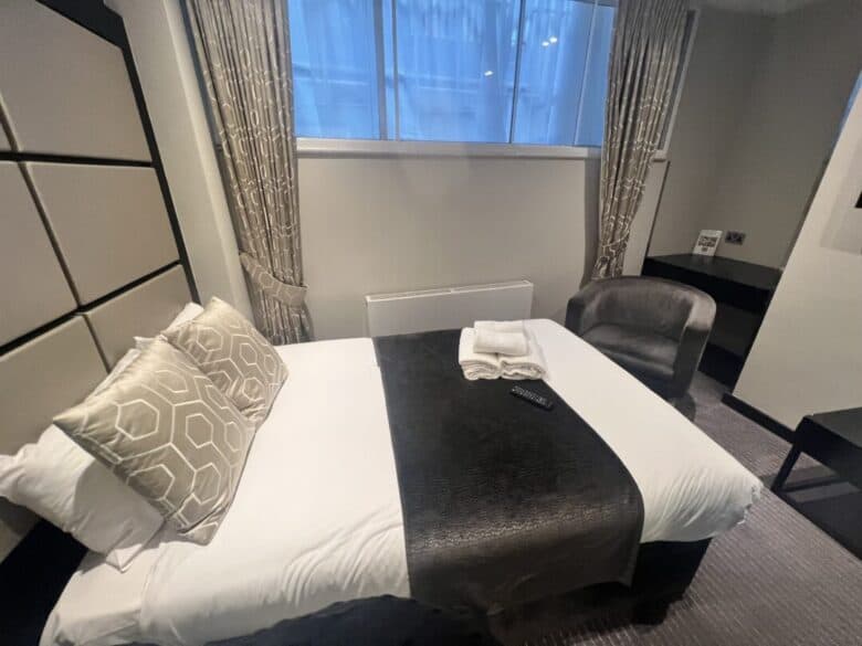 A cozy hotel room with a double bed, beige and patterned pillows, folded towels, and a remote on top. There's a gray armchair, curtained window, radiator, and a small black desk in the corner.