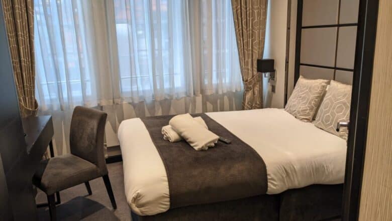 A neatly made hotel bed with white and brown bedding, two patterned pillows, and two folded towels. There is a chair by a dark desk, and large curtained windows let in natural light.