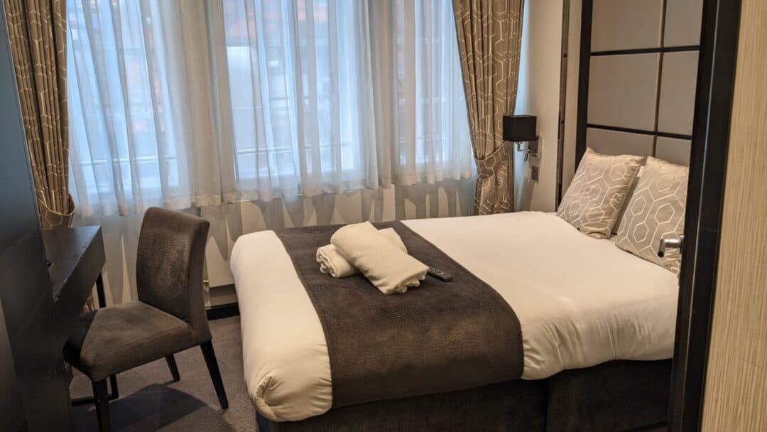A neatly made hotel bed with white and brown bedding, two patterned pillows, and two folded towels. There is a chair by a dark desk, and large curtained windows let in natural light.