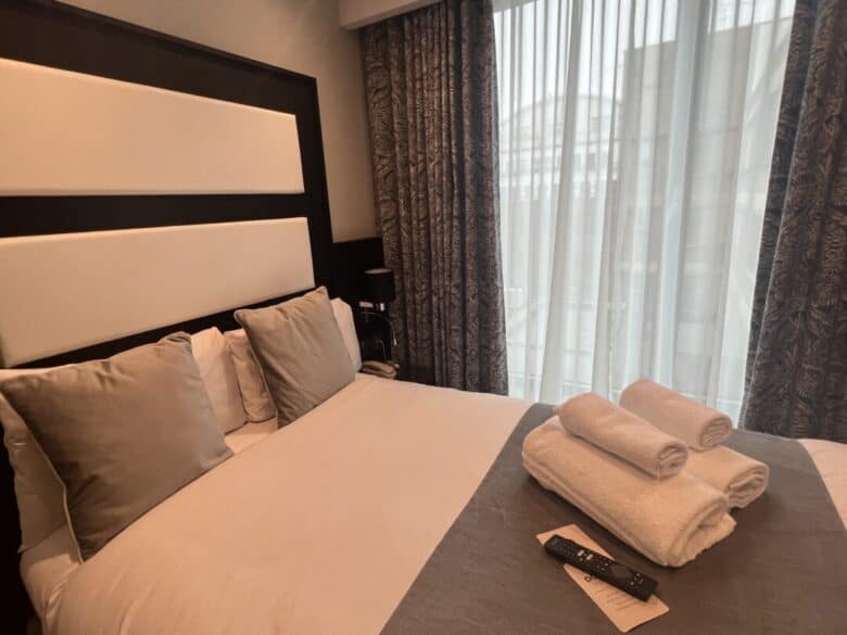 A neatly made hotel bed with four pillows, three rolled white towels, a remote control, and a note on top. Floor-to-ceiling windows with sheer curtains let in natural light.