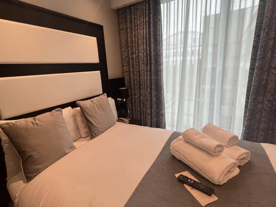 A neatly made hotel bed with four pillows, three rolled white towels, a remote control, and a note on top. Floor-to-ceiling windows with sheer curtains let in natural light.