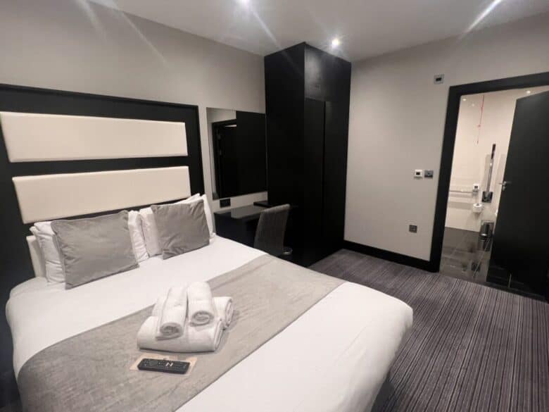 Modern hotel room with a double bed, gray and white bedding, towels placed on the bed, a wall-mounted TV remote, a desk with chair, large wardrobe, and an open door leading to a bathroom.