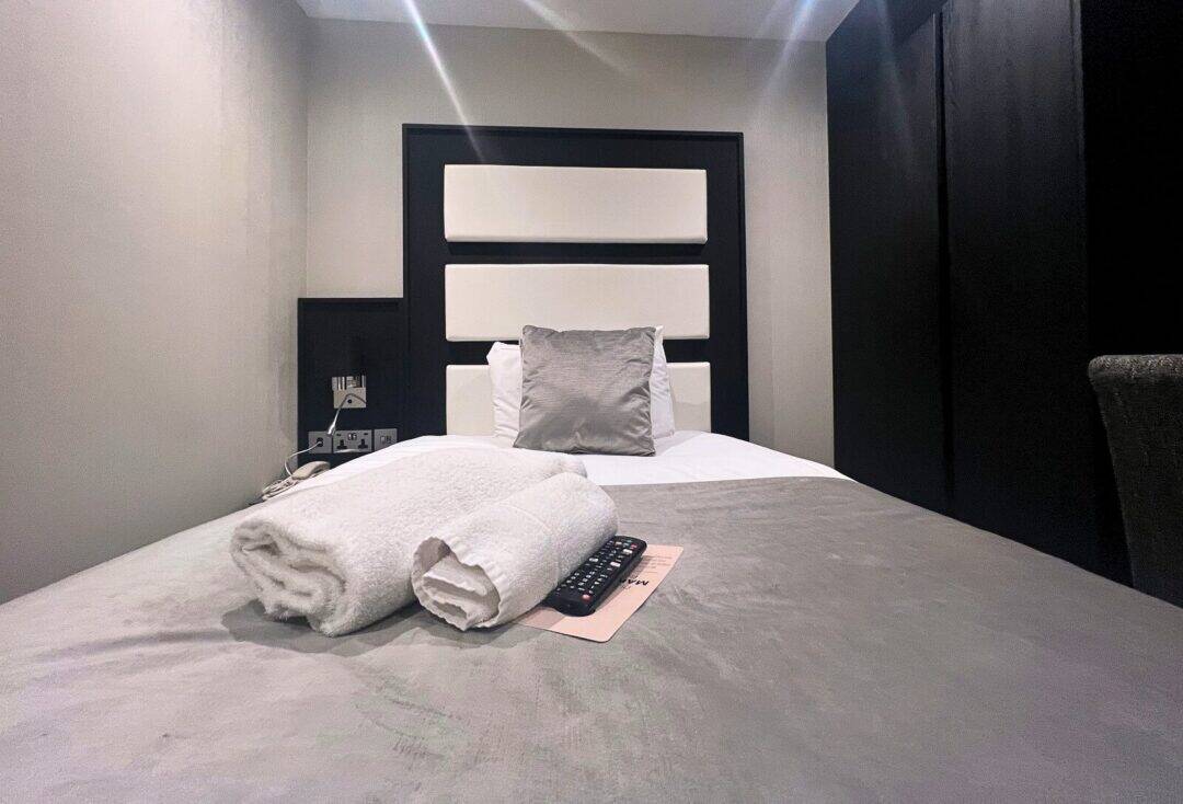 A neatly made single bed with a gray pillow and folded white towels on top, alongside a TV remote, in a modern, minimalistic hotel room with dark walls and a headboard.