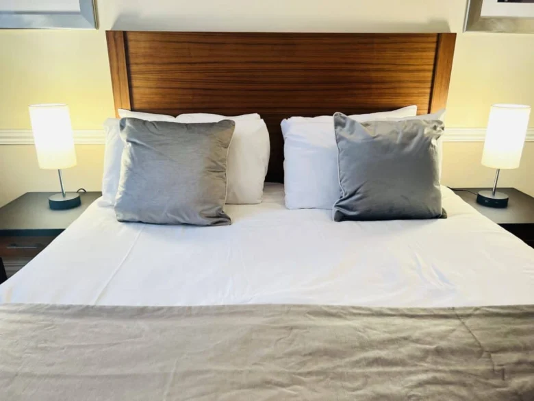 A neatly made bed with a wooden headboard, two gray throw pillows, white pillows, and a beige blanket. There are two small lamps on black nightstands on either side of the bed.