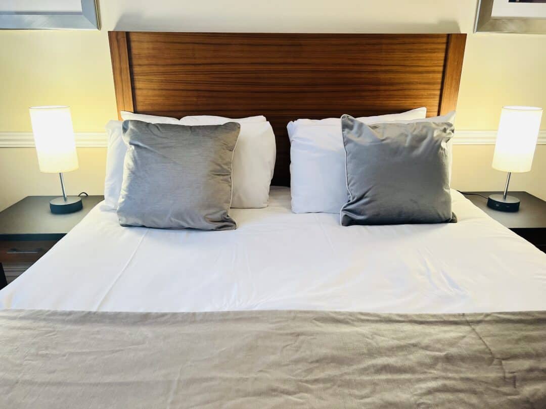 A neatly made bed with a wooden headboard, two gray throw pillows, white pillows, and a beige blanket. There are two small lamps on black nightstands on either side of the bed.